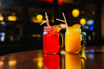 Two refreshing cold summer lemonades in glass jars, orange and red, served with ice, orange and lime slices. Shot in a bar using studio lights, reflections in a bar stand.