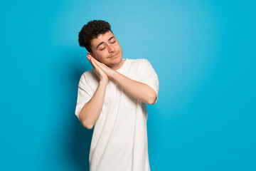 Young man over blue background making sleep gesture in dorable expression