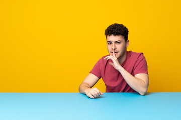 Young man with colorful wall and table showing a sign of silence gesture putting finger in mouth