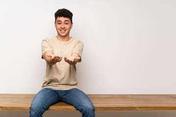 Young man sitting on table holding copyspace imaginary on the palm to insert an ad