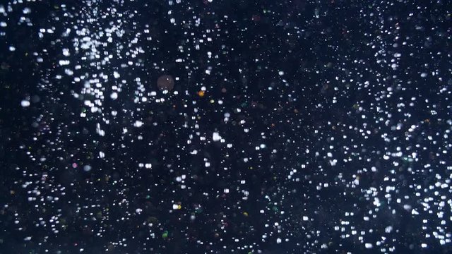 Glittering dark background suddenly filled out by numerous white snowball bokeh lights falling from above.