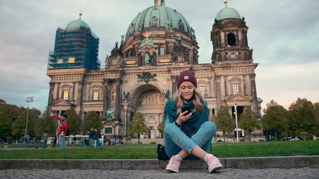 BERLIN, GERMANY - October 2018: Tourist girl in the autumn uses a mobile phone while sitting on the lawn near the cathedral in Berlin, Germany. Slow Motion.