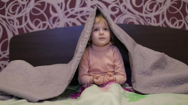 Little cheerful girl hides under blanket and watching TV. Concept of kids sleep