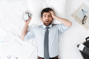 Shocked handsome young businessman in the morning in bed lies sleeping holding alarm clock.