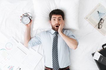 Shocked handsome young businessman in the morning in bed lies sleeping holding alarm clock.