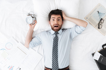 Shocked handsome young businessman in the morning in bed lies sleeping holding alarm clock.