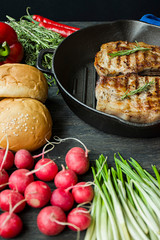 Grilled steak on a round grill pan, garnished with spices for meat, rosemary, greens and vegetables on a dark wooden background.