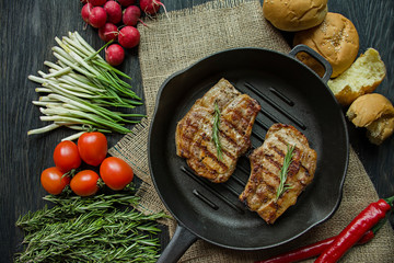 Grilled steak on a round grill pan, garnished with spices for meat, rosemary, greens and vegetables on a dark wooden background.