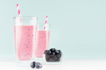 Blueberry milkshakes in transparent glass with blueberries in bowl, striped straw on white wood table and mint color wall, copy space.