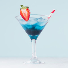 Summer blue alcohol cocktail with curacao  with strawberry red straw on light pastel green background and white wood table, square.