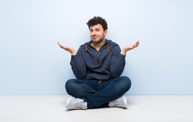 Young man sitting on the floor having doubts while raising hands