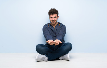 Young man sitting on the floor holding copyspace imaginary on the palm to insert an ad