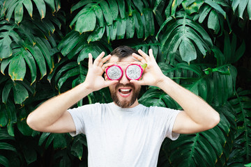 Young bearded man holding slices of Pitaya dragon fruit in front of his eyes, laughing out loud