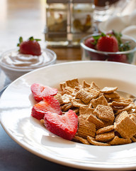 A bowl of cereal with strawberries as part of a balanced breakfast.  Yogurt, eggs, and strawberries blurred in the background.