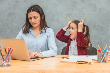 Daughter and mother on a gray background. During this, the girl puts two yellow crayons on her head, showing the tongue. Mom looks seriously while working on a laptop.