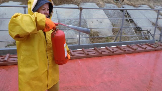 Elderly firefighter woman in a yellow raincoat puts on protective gloves and extinguishes the fire in the grill using fire extinguisher.