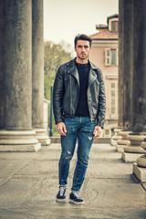 Obraz premium One handsome young man in urban setting in modern city, standing, wearing black leather jacket and jeans, looking at camera