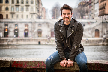 Fototapeta premium One handsome young man in urban setting in modern city, smiling, standing, wearing black leather jacket and jeans, looking at camera