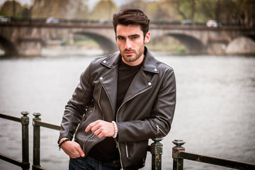 One handsome young man in urban setting in modern city, standing, wearing black leather jacket and jeans, looking at camera