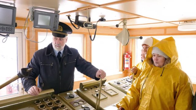 Elderly grey-haired captain of the Norway ship in the wheelhouse is talking with his team of sailors and workers in yellow raincoats and gives them instuctions.