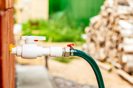 A rubber hose with a vertil for watering vegetation with water comes out of the wall of a wooden house, on a blurred fence and firewood.