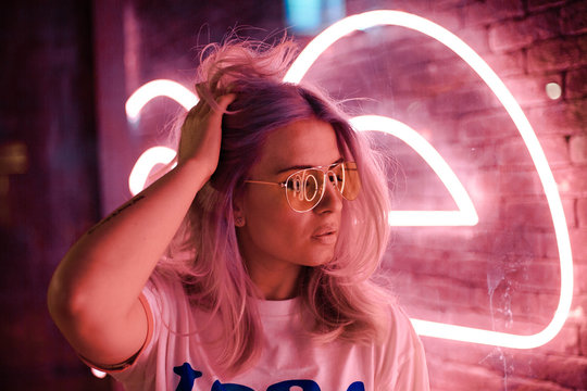 Lovely Blonde Woman With Glasses On The Street With Neon Background