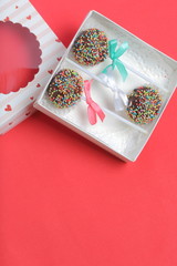 Cake Pops in chocolate with colored sprinkles. Decorated with a ribbon bow. They lie in a gift box, in the lid of which there is a transparent window.  On a red background.