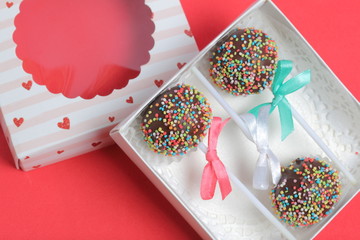 Cake Pops in chocolate with colored sprinkles. Decorated with a ribbon bow. They lie in a gift box, in the lid of which there is a transparent window.  On a red background.