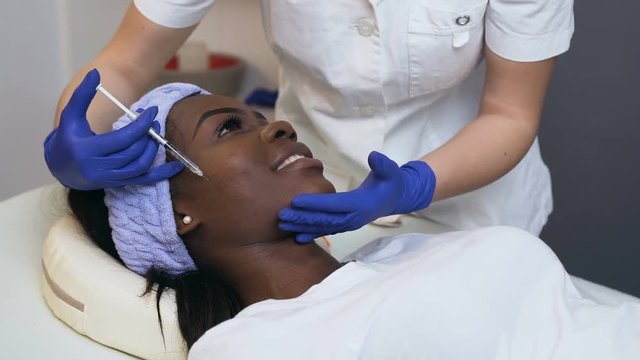 Side view of female hands of cosmetologist making beauty injection for young african woman.