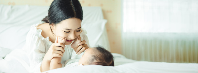 Panoramic banner image of side view of a young Asian mother hands holding newborn baby feet on a white bed. Maternity concept, soft image of beautiful family, mother's day concept. wide crop - obrazy, fototapety, plakaty