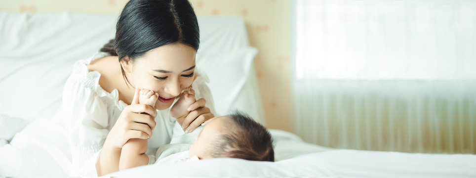 Panoramic banner image of side view of a young Asian mother hands holding newborn baby feet on a white bed. Maternity concept, soft image of beautiful family, mother's day concept. wide crop