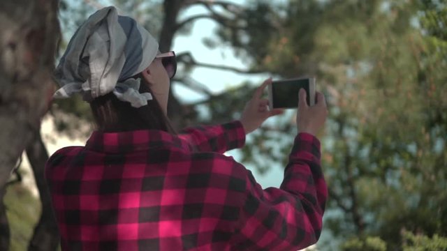 A girl with dark hair in a black and red shirt, a bandana and glasses stands between the trees, makes a photo on the phone. Background mountains, sky.