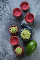 Freshly made guacamole sauce with red vegetable tartlets on a grey concrete background, flatlay, vertical shot