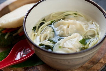 Close-up of wonton soup with egg noodles served in a bowl with gua bao in the background, studio shot