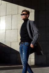 Fashionable young adult man in sunglasses. Posing in grey elegant jacket, black polo neck, jeans trousers and brown mocassin shoes. Fashion trends.