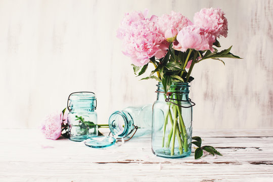 Beautiful pink Peony flowers in an antique blue mason jar over a white rustic wood table background  with copy space for your text. Image edited to give farmhouse decor style.
