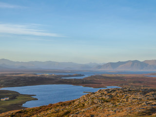 A flatland, covered with golden grass. Dried grass due to the winter season. Fjord view, with some taller mountains in the back. Calm water of the fjord. Clear weather with some clouds on the sky.