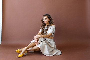European young brunette woman sitting in studio and posing, brown background. In full growth.