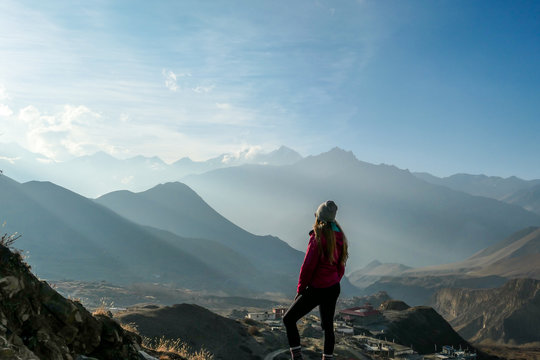 A girl wearing pink jacket standing on a top of a mountain and enjoying the misty Himalaya range spreading in front of her. The sun light is nicely marked. Freedom and achievement.