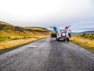 A couple stretching from the sides of the car and waving at the camera. The car is pulled on the side of the road, next to mountains. Mountains are overgrown with yellow and green grass. Fun moments © Chris