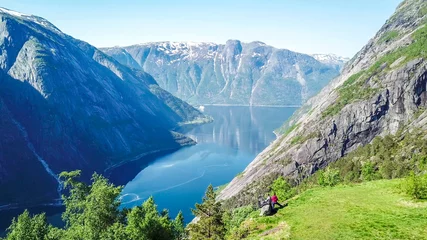 Fotobehang A couple standing on the meadow with a majestic view on Eidfjord from Kjeasen, Norway. Slopes of the mountains are overgrown with lush green grass. Water has dark blue color. Sunny and clear day © Chris