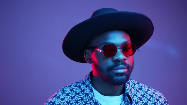 Attractive hot Afro American hipster man in stylish hat takes off his trendy steampunk polarized eyeglasses and seductively smiles straight to camera on violet blank background in bright neon lights