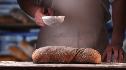 In the bakery, a baker after baking fresh bread sprinkles sugar powder (flour) on bread (flour...