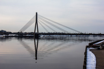 Fototapeta na wymiar Huge suspension bridge and its refliction in a wide river Daugava during winter period in Riga, Latvia.