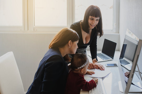 close up photo of two women in office outfit working on computers and discussing a report on the floor; one of them is with a baby girl in her arms