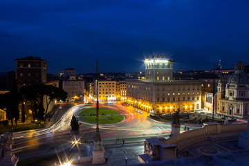 Fototapeta premium Square in the center of Rome in Italy at night with colorful cars tracks, trails. Long exposure image. Historical and residential buildings on the background and statue of palace on the foreground.
