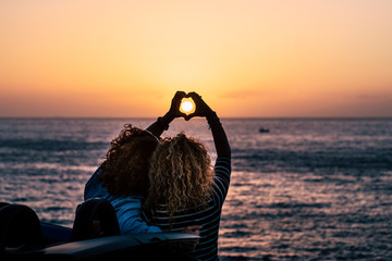 Romantic friendship people concept with two curly lady viewed from back doing hearth love sign with hands to celebrate the summer holiday vacation travel in front of the beauty of nature ocean