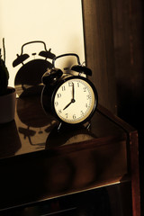 Black retro vintage alarm clock on a wooden bedside table with reflection in a polished surface in the rays of the setting sun with hard shadows