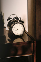 Black retro vintage alarm clock on a wooden bedside table with reflection in a polished surface in the rays of the setting sun with hard shadows