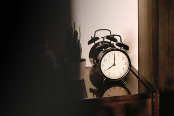 Black retro vintage alarm clock on a wooden bedside table with reflection in a polished surface in the rays of the setting sun with hard shadows